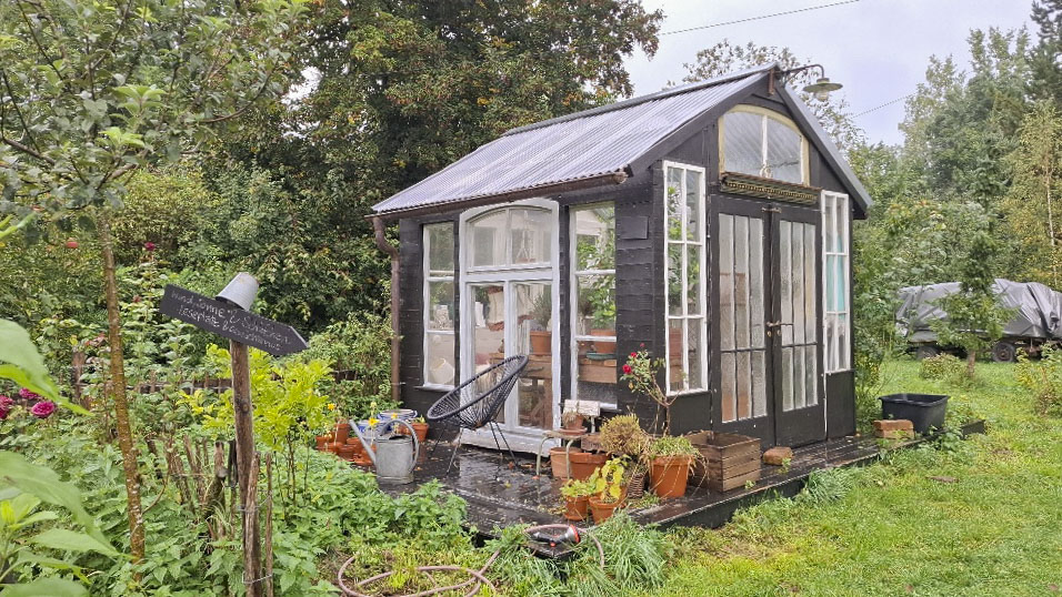 Schwarze Gartenhütte mit großen Sprossenfenstern und Topfpflanzen im üppigen Garten