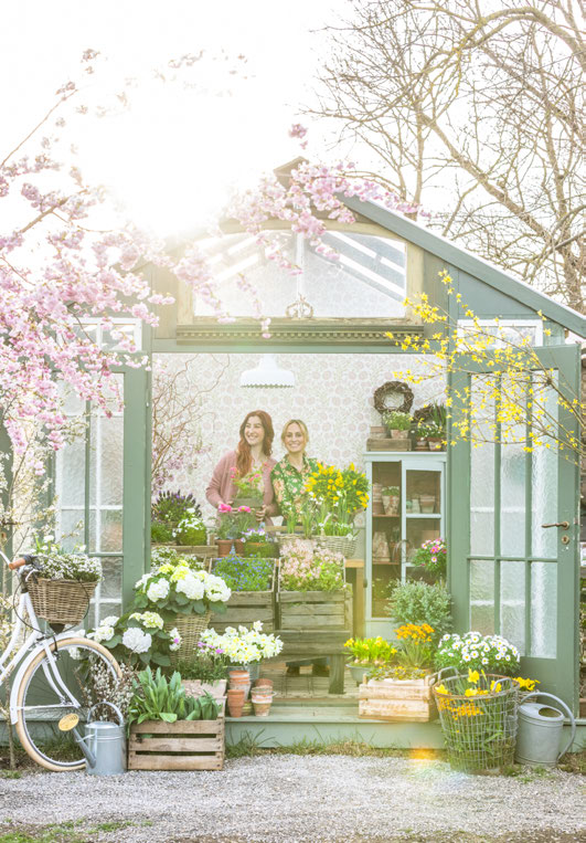 Mintgrünes Gewächshaus vor blühenden Kirschen, davor zwei Frauen, Frühlingsblumen und Korbtaschen