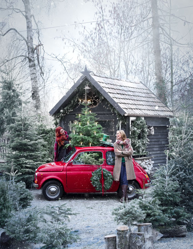 Roter Fiat 500 mit Weihnachtskranz und kleinem Tannenbaum auf dem Dach vor schwarzer Hütte im verschneiten Wald